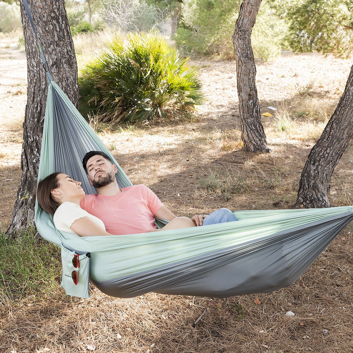 Couple relaxing in a double camping hammock tied between two trees in a sunny outdoor setting, ideal for hiking and camping.