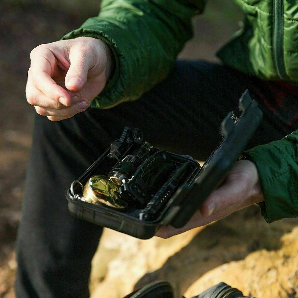 Person outdoors holding an open 14-in-1 survival kit with tactical tools and compass in a rugged case.